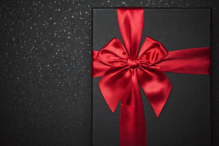 Black gift box with a red ribbon and a large bow on a granite dark background. Close-up photo. Place...