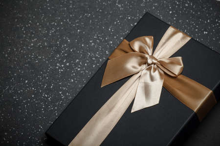 Black gift box with a golden ribbon and a large bow on a granite dark background. Place for the...