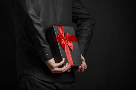 Man in a black shirt holds a gift with a red ribbon behind his back. Black gift box in hands....