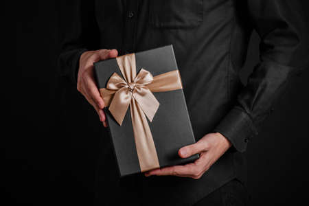 A guy in a black shirt is holding a gift with a goldeb ribbon. Black background. Black gift box in hands.の写真素材