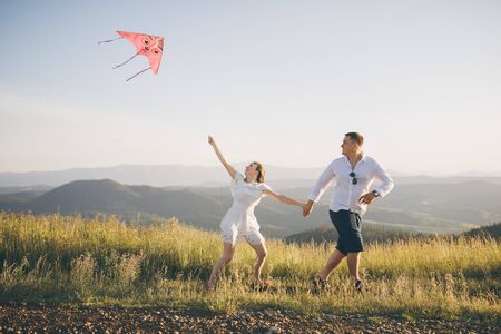 Running with flying kite have fun on countryside. Boy and girl running on a hill with a flying kite against a blue sky and sun rays.の写真素材