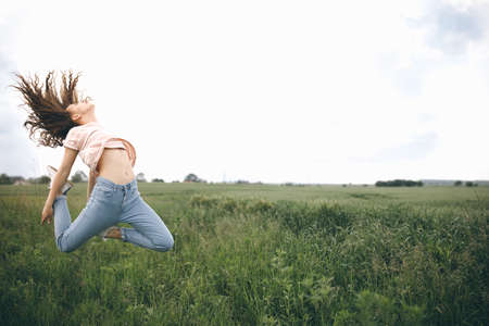 A Young Happy Girl With Curly Hair Jumps On The Green Field Against Cloudy Sky.