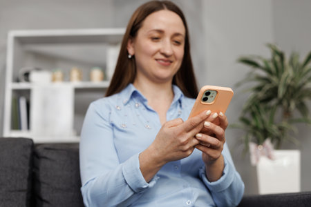Happy casual beautiful woman talking on phone while sitting on sofa at home. Smiling young woman using mobile phone at homeの写真素材