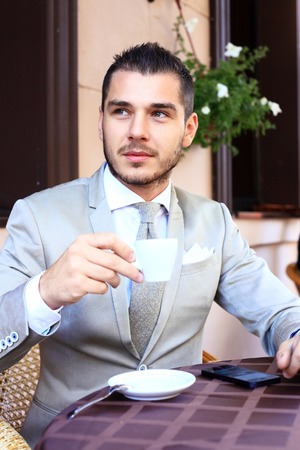 Young businessman drinking a cup of coffee while sitting at a coffee shop terrace table, outdoors and smiling at the cameraの写真素材