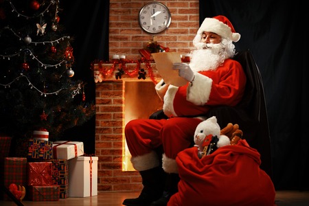 Portrait of happy Santa Claus sitting at his room at home near Christmas tree and big sack and reading Christmas letter or wish listの写真素材