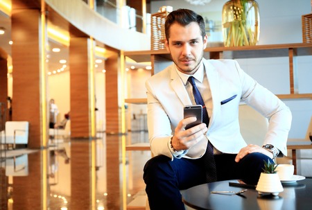 Happy young businessman sitting relaxed on sofa at hotel lobby using smartphone, waiting for someone.の写真素材