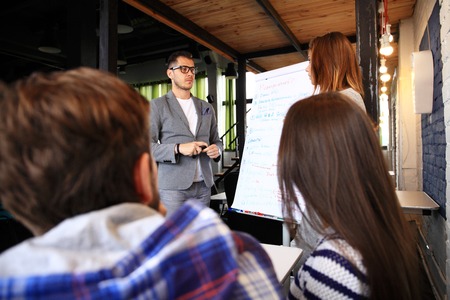 Take a look at our results! Handsome young man in glasses standing near whiteboard and pointing on the chart while his coworkers listening and sitting at the tableの写真素材