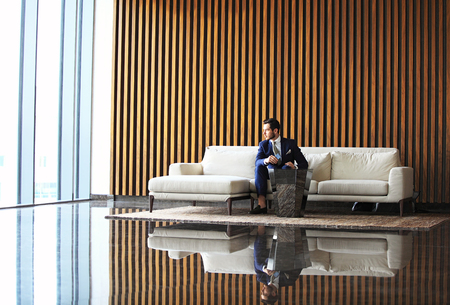 Happy young businessman sitting relaxed on sofa at hotel lobby, waiting for someoneの写真素材