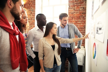 Collaboration is a key to best results. Group of young modern people in smart casual wear planning business strategy while young woman pointing at infographic displayed on white wall in the officeの写真素材