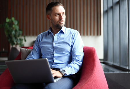 Smiling young handsome businessman holding cup of coffee and working with laptop in cafe indoorsの写真素材