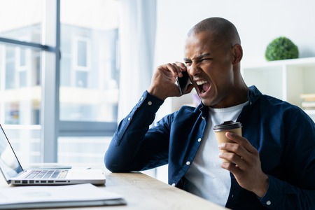 Young man talking on his mobile phone in office. African executive sitting at his desk with laptopの写真素材
