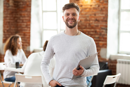 Confident team leader. Confident young man keeping arms crossed and looking at camera with smile while his colleagues working in the background.の写真素材