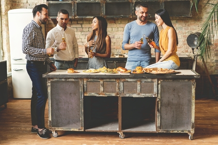 Home party. Full length of cheerful young people enjoying home party while communicating and eating snacks on the kitchen.の写真素材