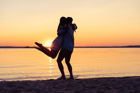 Silhouette of happy couple on beach at sunset, man taking the girl in his arms.