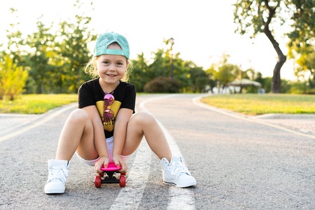 Fashion little girl child sitting on skateboard in city, wearing a sunglasses and t-shirt.