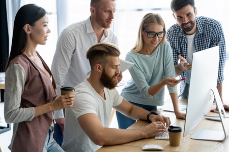 Discussing successful project. Group of young cheerful business people working and communicating while sitting at the office desk together with colleagues sitting in the background.の写真素材