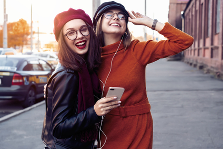 Two joyful young girls dancing while listening to music on smartphone, city outdoor.の写真素材