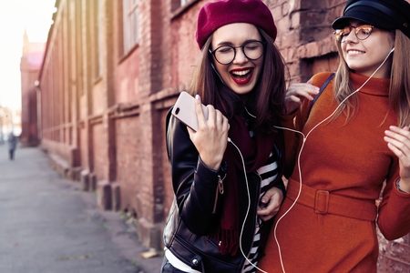 Two joyful young girls dancing while listening to music on smartphone, city outdoor.の写真素材
