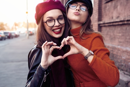 Outdoors fashion portrait young pretty best girls friends in friendly hug. Walking at the city. Posing at the street. Wearing stylish outerwear and hats. Bright make up. Positive emotions.の写真素材