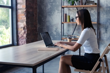 Concentrated at work. Confident young woman in smart casual wear working on laptop while sitting near window in creative office or cafe.の写真素材