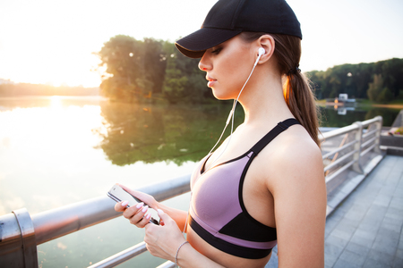 Tired fitness woman sweating taking a break listening to music on phone after difficult training. Young woman listening to music with earphones on smart phone app for fitness motivation.の写真素材