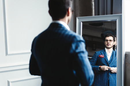 Perfect look. Reflection of handsome young man in full suit adjusting his jacket while standing in front of the mirror indoors.の写真素材