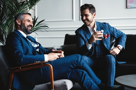 Relaxing. Full length of two young handsome men in suits holding glasses and looking at each other while resting indoors.の写真素材