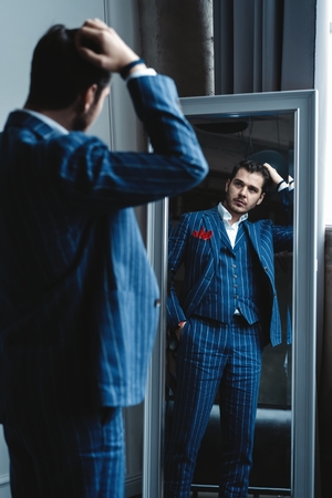 Perfect look. Reflection of handsome young man in full suit adjusting his jacket while standing in front of the mirror indoors.の写真素材