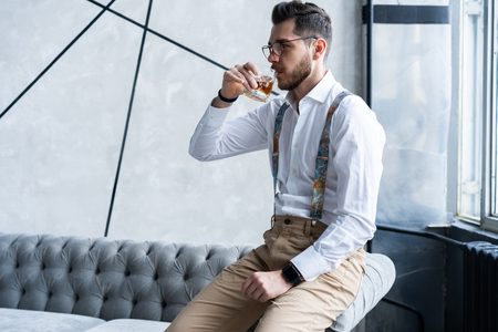 Feeling lonely. Stylishly dressed young man holding a glass while sitting on sofa.の写真素材