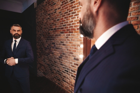 Elegant look. Full length of handsome man in full suit adjusting his jacket while standing in front of the mirror indoorsの写真素材