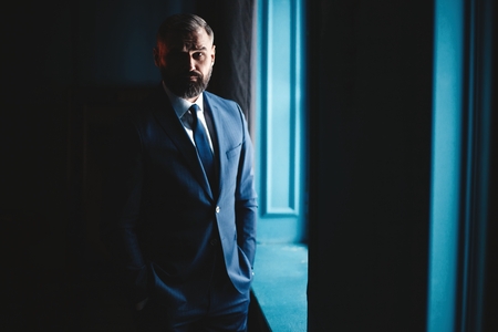 Thinking about... Thoughtful man in full suit looking away through the window while standing indoors.の写真素材