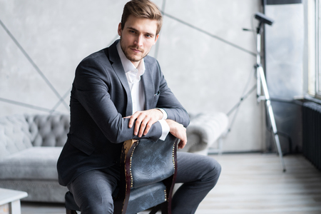 Portrait of confidence. Thoughtful young man in full suit looking away while sitting on the stool.の写真素材