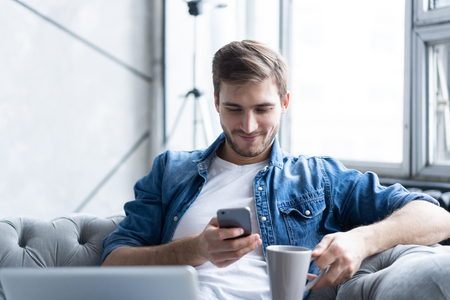 Young man using his smartphone for online banking - sitting on sofa with laptop on leap.の写真素材