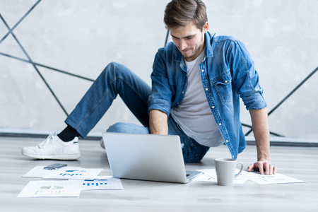 Man working at home. Handsome young man sitting on the floor and examining document while laptop and documents laying near him.の写真素材
