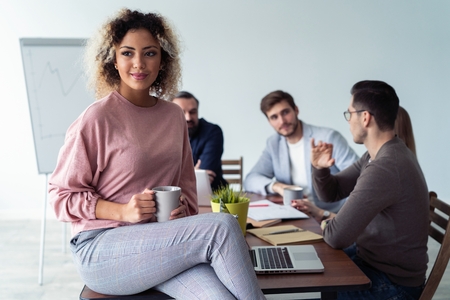 Happy to be in team. Beautiful cheerful African woman looking at camera with smile while sitting at the office table with her coworkers.の写真素材