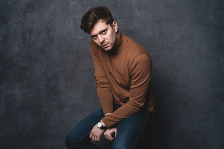 handsome casual man sits on chair near a dark gray wall and looks to side, portrait picture.