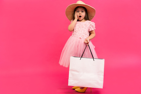 Happy lovely moments of shopping time with cute little girl in dress standing in mother`s big shoes with white packages in hands isolated on pink backgroundの写真素材