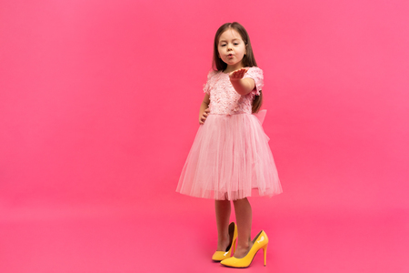 Fashionable cute young girl with long brunette hair, in stylish dress wearing big mother`s shoes, sending kiss to camera isolated on pink background. Having fun of joyful kid. Place for text.の写真素材