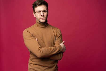 Smiling Man in glasses and sweater standing with crossed arms in studio and looking away. Isolated pink background.の写真素材