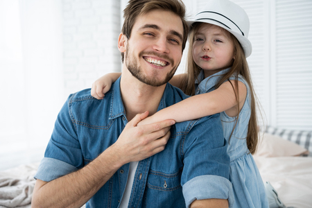 Portrait of handsome father and his cute daughter hugging, looking at camera and smiling while sitting on sofa at home.の写真素材