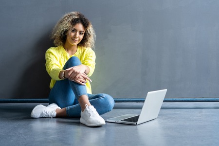 Happy young woman sitting on the floor with crossed legs and using laptop on gray background.
