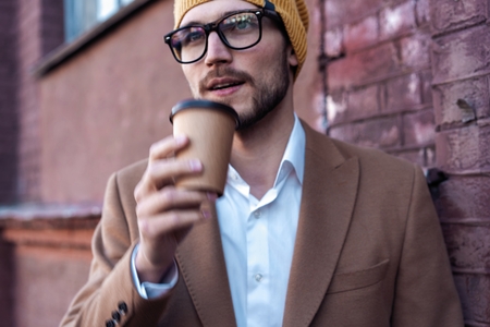 Handsome young man in casual wear holding disposable cup and smiling while walking through the city street.の写真素材