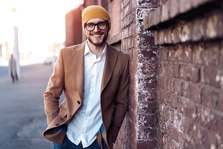 Portrait of stylish handsome young man in glasses with bristle standing outdoors. Man wearing jacket and shirt, leaning against wall.の写真素材