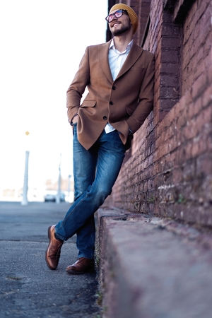 Portrait of stylish handsome young man in glasses with bristle standing outdoors. Man wearing jacket and shirt, leaning against wall.の写真素材