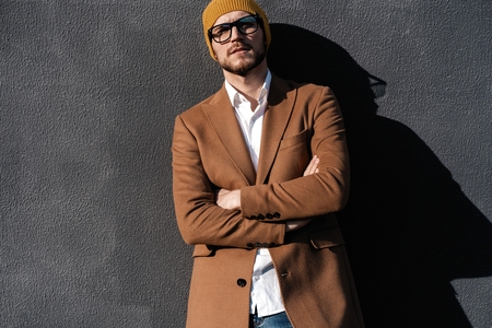 Portrait of stylish handsome young man in glasses with bristle standing outdoors. Man wearing jacket and shirt, leaning against wall.の写真素材