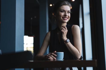 Portrait of gorgeous female drinking tea or coffee and looking with smile out of the coffee shop window while enjoying her leisure timeの写真素材