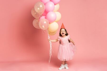 Happy celebration of birthday party with flying balloons of charming cute little girl in tulle dress smiling to camera isolated on pink background. Charming smile, expressing happinessの写真素材