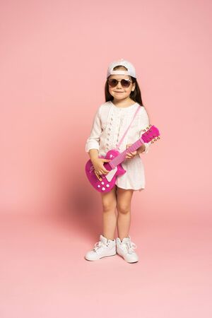 Laughing fashion little girl having fun with guitar isolated on pink background.の写真素材