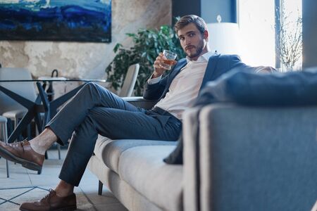 Handsome and successful businessman in stylish suit holding glass whiskey while sitting at office.