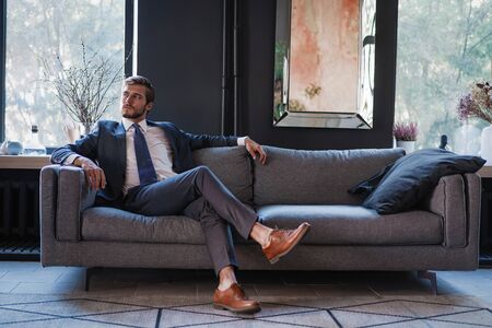 Young and charming businessman is thinking about business while sitting on the sofa in his modern office.の写真素材